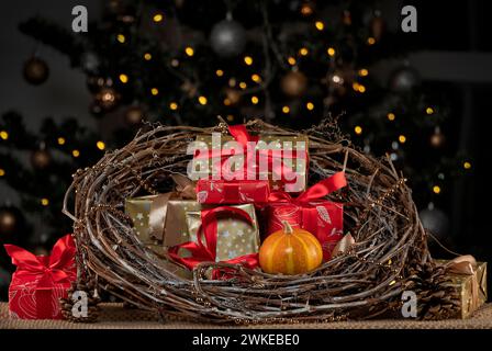Weihnachtlicher gewebter Kranz mit Geschenken im Inneren auf einem Hintergrund eines verschwommenen, verzierten Weihnachtsbaums. Stockfoto