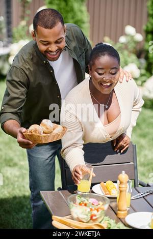 Glückliches afroamerikanisches verheiratetes Paar serviert Tisch im Garten, Brötchen und ein Glas Orangensaft Stockfoto