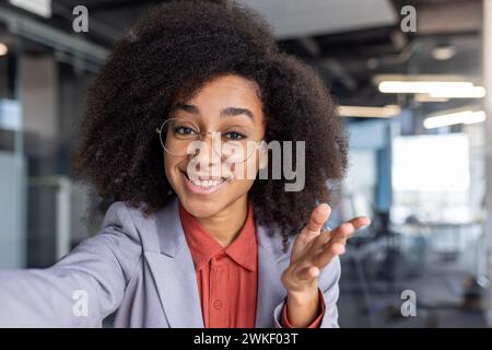 First-Person-Ansicht einer schwarzen Frau im Business-Anzug, die mit einem Zahnlächeln in die Kamera schaut und mit der Hand Gesten zeigt. Moderner smm Manager sendet Videos aus dem Bürogebäude, um die Bekanntheit zu erhöhen. Stockfoto