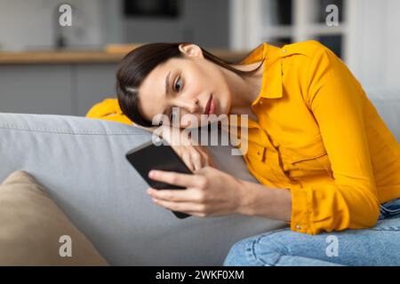 Besorgte junge Frau, die auf der Couch liegt und auf das Telefon blickt Stockfoto