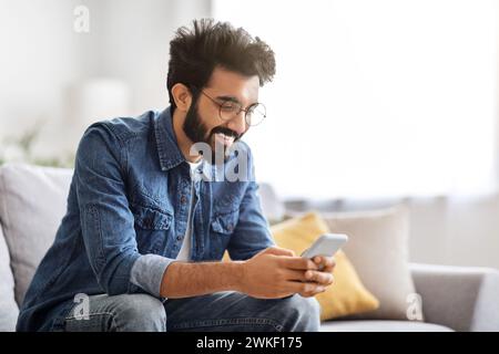 Fröhlicher bärtiger indianer, der zu Hause mit einem Lächeln auf sein Smartphone schaut Stockfoto
