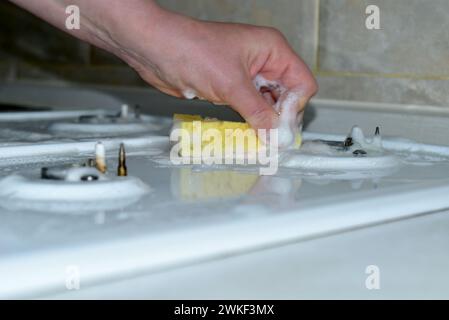 Nahaufnahme einer Frauenhand, die die Oberfläche eines Gasherds um einen kleinen Brenner mit einem Schwamm und Waschmittel wäscht. Stockfoto