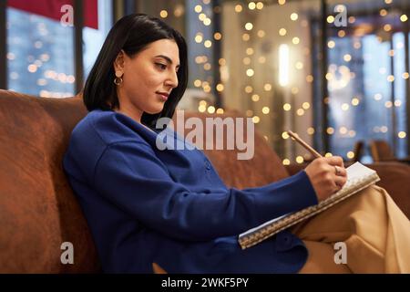 Seitliches Porträt einer jungen Frau aus dem Nahen Osten, die in Notizbuch oder Zeichnung schreibt, während sie im Café auf der Couch sitzt Stockfoto