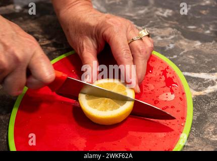 Frauenhände teilen eine gelbe Zitronenscheibe in zwei Hälften, während sie das Messer halten und die Frucht mit den Fingern stabilisieren. Enthält ein rotes Schneidebrett. Stockfoto