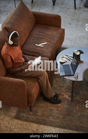 Vertikales Hochwinkelporträt kreativer Schwarzer Mann, der Skizzen auf der Couch im Café trägt und Kopfhörer trägt Stockfoto