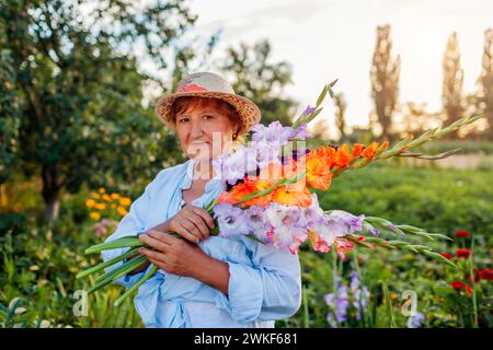 Porträt eines glücklichen Gärtners, der frische Gladiolusblüten hält, die im Sommergarten geerntet wurden. Die Seniorin pflückte Blüten, die organisch gewachsen sind Stockfoto
