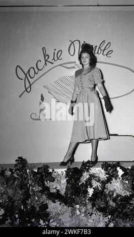 1950er Jahre, historisch, eine elegante junge Dame posiert auf einer Bühne in einem langen wadenlangen, modischen Jackie-flinken Kleid + Handschuhe und Hut. Stockfoto