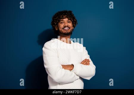 Porträt des jungen gutaussehenden lockigen Haares indischer positiver Mann im weißen Pullover gefaltete Hände lächelnd isoliert über dunkelblauem Hintergrund Stockfoto