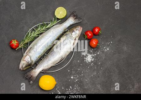 Rohe Fischforelle mit Rosmarinzweigen, Zitronenscheiben und Tomaten. Flache Lagen. Kopierbereich. Schwarzer Hintergrund Stockfoto