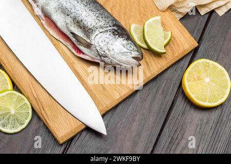 Geschälte Fischforelle und Messer auf Schneidebrett. Zitronenscheiben auf dem Tisch. Dunkler Holzhintergrund. Flache Lagen Stockfoto