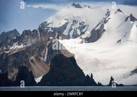 Ein großer Aufschluss von Basaltformationen, der aus einer Bucht vor dem Hintergrund von hohen Berggipfeln und einem Gletscher aufsteigt. Antarktische Halbinsel, Antarktis Stockfoto