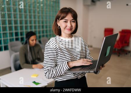 Porträt einer freundlichen jungen Geschäftsfrau in lässiger Kleidung, die den Laptop in der Hand hält, im Büro steht und lächelnd in die Kamera blickt. Vielseitiges Geschäftsteam Stockfoto