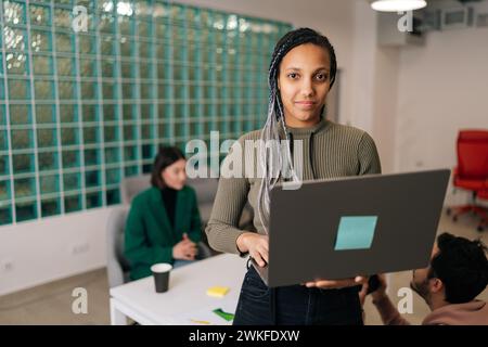 Porträt einer jungen afroamerikanischen Geschäftsfrau, die im modernen Büro steht und einen Laptop hält und in die Kamera blickt. Stockfoto