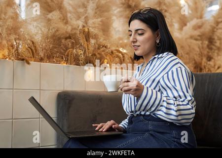 Seitliches Porträt einer eleganten Frau aus dem Nahen Osten mit Laptop, der in der Bürolounge auf der Couch sitzt und eine Kaffeetasse hält Stockfoto