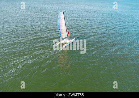 Drohnenansicht eines Katamarans, das im türkisfarbenen Wasser des El Portil Beach mit einem mehrfarbigen Segel in Punta Umbria Municipali segelt Stockfoto