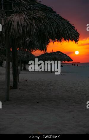 Eine Reihe von strohgedeckten Sonnenschirmen am Strand bei Sonnenuntergang, die Schatten spenden, während Himmel und Wasser sich in eine malerische Szene der Abenddämmerung verschmelzen Stockfoto