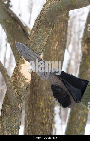 Nahaufnahme der menschlichen Hand in schwarzem Handschuh, der ein taktisches Messer hält und im Winter einen Holzstock schneidet. Handmesser. Outdoor Survival Activities Konzept. Stockfoto