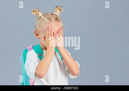 Zurück zur Schule. Kleines Schulmädchen mit Rucksack, der ihr Gesicht mit den Händen bedeckt Stockfoto