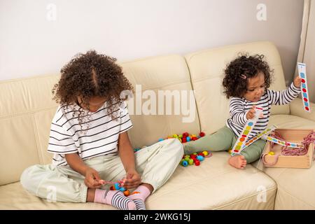 Ein junges afrikanisch-amerikanisches Mädchen und ein Kleinkind sitzen auf einem Sofa, beide mit pädagogischem Spielzeug besetzt. Pädagogisches Spiel, Beruf Stockfoto