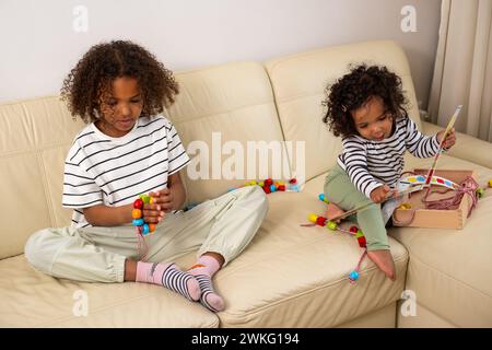 Zwei afroamerikanische Kinder spielen auf einem cremefarbenen Sofa mit pädagogischem Spielzeug. Die ruhige Atmosphäre bedeutet, dass man lernen muss Stockfoto