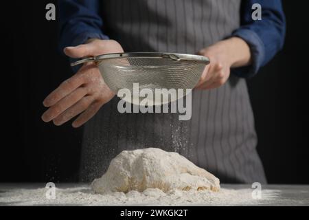 Brot machen. Frau siebt Mehl über Teig am Tisch auf dunklem Hintergrund, Nahaufnahme Stockfoto