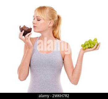 Frau, Versuchung und Dessert über Obst im Studio mit weißem Hintergrund im Hintergrund. Mädchen, Wahl und Entscheidung zwischen Muffin oder Trauben zur Ernährung Stockfoto