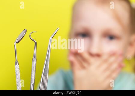 Zahninstrumente vor dem Hintergrund eines verängstigten Mädchens, das ihren Mund mit den Händen bedeckt. Das Konzept der Angst vor Zahnärzten bei Kindern. Stockfoto