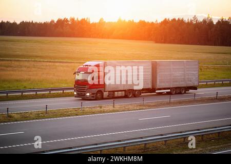 Ein Lkw mit Auflieger und Anhängevorrichtung transportiert Ladung vor dem Hintergrund eines Sonnenuntergangs entlang der Autobahn. Massengütertransport Konzept, Logist Stockfoto