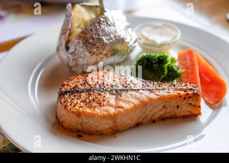 Mit Pfeffer gewürzter Lachs wird mit einer in Alufolie gebackenen Kartoffel, Brokkoli, Karotten und cremiger Sauce auf einem weißen Teller in Norwegen serviert. Stockfoto