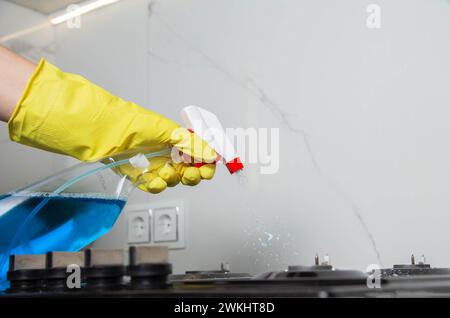 Die Hand eines Mädchens mit einem Reinigungsspray sprüht auf einen Gasherd. Modernes fettfreies, antistatisches Reinigungsmittel. Hintergrund: Weiße Porzellanfliesen Stockfoto