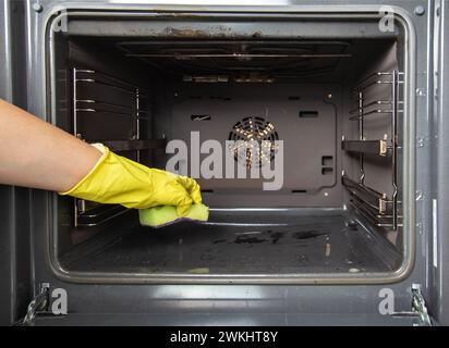 Ein Mädchen hält einen Schwamm in der Hand vor dem Hintergrund eines Ofens. Konzept zur Reinigung von Elektroöfen in der Küche. Reinigungsfirma Stockfoto