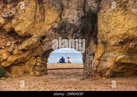 Gehen Sie unter die Klippe am Strand Praia da Batata mit einem Paar, das auf Sand sitzt und auf das Meer in Lagos, Algarve, Portugal blickt. Stockfoto