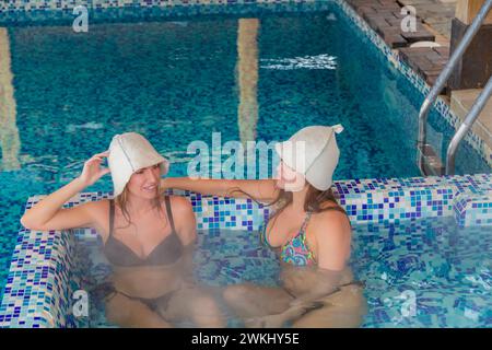 Zwei Frauen tragen Badeschutzhüte und entspannen sich in einem Whirlpool am Pool, genießen Freizeit und Spaß Stockfoto