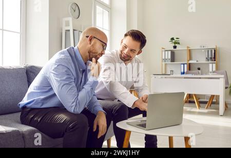 Zwei männliche Kollegen, die im Büro zusammen mit dem Laptop sitzen Stockfoto
