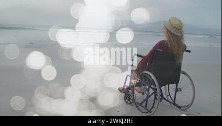 Bild von Lichtflecken über behinderten kuakasischen Frauen, die im Rollstuhl am Strand sitzen Stockfoto