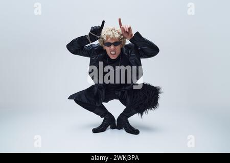Junger Mann in schwarzer Kleidung und Stiefeln, rocken Gesten, mit einer lebhaften und rebellischen Haltung vor grauem Studiohintergrund. Stockfoto