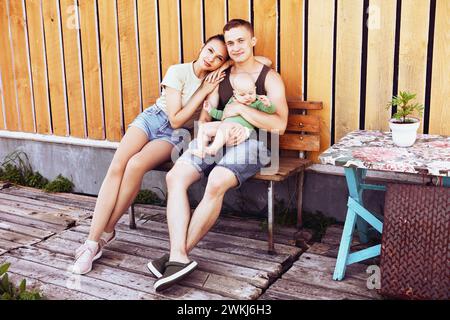 Junge Eltern mit Baby in den Armen sitzen im Sommer im Hinterhof ihres Hauses, Mann und Frau sind Anfang 20, in T-Shirts und Shorts gekleidet. Stockfoto