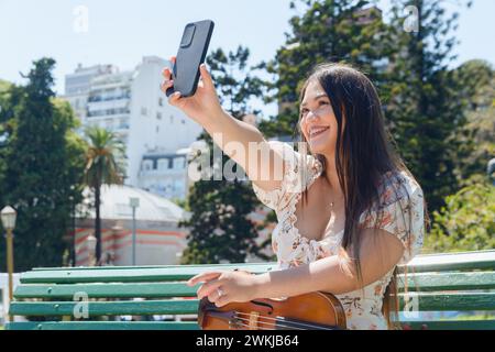 Die junge Latina Buskergeigerin sitzt mit ihrer Geige draußen auf der Bank, auf der plaza, lächelt, macht Selfie mit ihrem Handy, Technologie-Conce Stockfoto