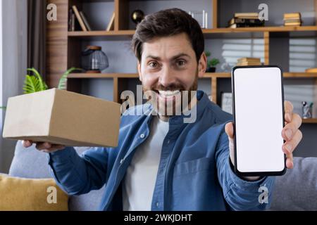 Glücklicher Mann, der ein Paket hält und einen leeren Telefonbildschirm zeigt, ideal für Anzeigen und App-Mockups, mit einer gemütlichen Heimumgebung im Hintergrund. Stockfoto