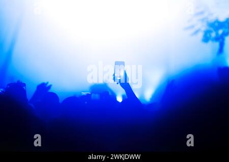Silhouette einer Hand, die ein Smartphone hält, um einen Moment während eines Nachtkonzerts festzuhalten, das von Bühnenlichtern beleuchtet wird Stockfoto