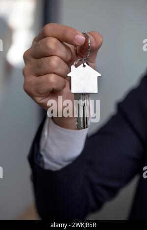 Das Bild zeigt eine Personenhand, die einen silbernen Hausschlüssel mit einem hausförmigen Schlüsselanhänger hält. Der Schlüssel ist auf weißem Hintergrund isoliert. Stockfoto