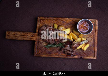 Zwei saftige Lammkoteletts und knusprige Kartoffelscheiben serviert auf einem rustikalen Holzbrett mit einer Beilage aus würziger Dippsoße. Stockfoto