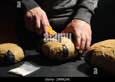 Schmuggel und Drogenhandel. Mann, der ein Paket mit Betäubungsmitteln öffnet, mit einem Cutter am dunklen Tisch, Nahaufnahme Stockfoto