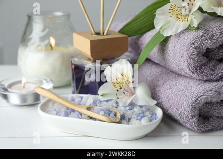 Spa-Zusammensetzung. Handtücher, brennende Kerzen, Schilflufterfrischer, Meersalz und schöne Blumen auf weißem Holztisch, Nahaufnahme Stockfoto