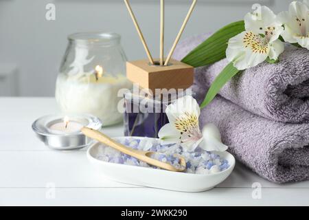 Spa-Zusammensetzung. Handtücher, brennende Kerzen, Schilflufterfrischer, Meersalz und schöne Blumen auf weißem Holztisch, Nahaufnahme Stockfoto