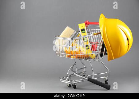 Kleiner Einkaufswagen mit Bauhöhe, Bürste, Handschuhe und Schutzhelm auf grauem Hintergrund. Leerzeichen für Text Stockfoto