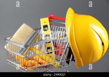 Kleiner Einkaufswagen mit Bauhöhe, Bürste, Handschuhe und Schutzhelm auf grauem Hintergrund Stockfoto