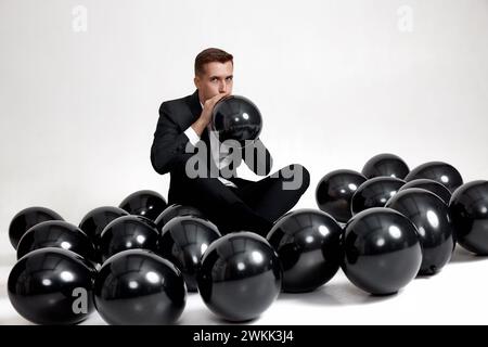 Schöner Mann in schwarzer Jacke, der schwarzen Luftballon auf weißem Studiohintergrund bläst. Neujahrsfeier Stockfoto