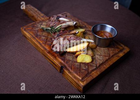 Zwei saftige Lammkoteletts und knusprige Kartoffelscheiben serviert auf einem rustikalen Holzbrett mit einer Beilage aus würziger Dippsoße. Stockfoto