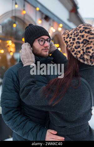 Glückliches, wunderschönes Paar von Liebhabern in modischer Winterkleidung mit Jacke auf der Straße. Das hübsche Mädchen passt den Hut ihres Freundes an Stockfoto